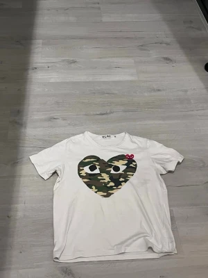 Vit t-shirt med camo hjärta - Säljer en vit t-shirt med ett stort hjärta i camouflage-mönster och ögon på bröstet. Hjärtdesignen har även en liten röd detalj. T-shirten har rund hals och korta ärmar. Perfekt för dig som gillar streetwear och coola prints.