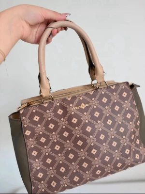 Brun mönstrad handväska från Vizzano - Snygg handväska från Vizzano i brunt och beige med geometriskt rutmönster. Väskan har två stabila handtag i beige konstläder och guldfärgade metalldetaljer. Sidorna är i mörkare brunt material och väskan stängs med dragkedja.