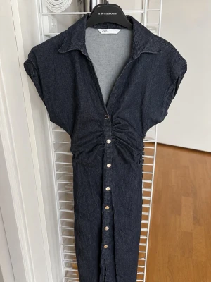 Mörkblå jeansklänning med knappar - Snygg mörkblå jeansklänning från ZARA med korta ärmar, krage och knappar framtill. Klänningen har en markerad midja och lätt rynk vid bysten för en cool look. Perfekt till sneakers eller boots.