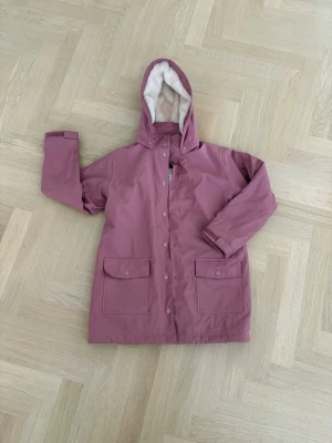 Rosa regnjacka från KappAhl - Mysig rosa regnkappa från KappAhl med fluffigt vitt foder och stor huva. Jackan har två stora fickor framtill, tryckknappar och är tillverkad i ett vattenavvisande material. Perfekt för kalla dagar och ger en trendig look.