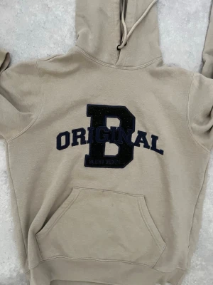 Beige hoodie med tryck 'B ORIGINAL' - Säljer en beige hoodie med stor svart bokstav 'B' och texten 'ORIGINAL' i mörkblått på bröstet. Har huva med dragsko och en klassisk magficka. Perfekt för en chill och avslappnad stil.