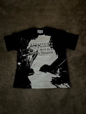 Maison Margiela svartvit t-shirt - Maison Margiela t-shirt i svart och vitt med abstrakt tryck framtill och logotyp samt siffror. Klassisk passform och korta ärmar. Perfekt för dig som gillar statement-plagg och streetwear-vibe.