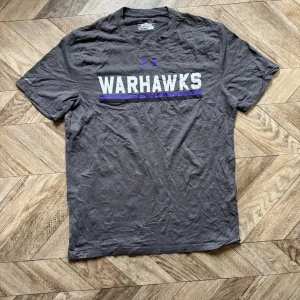 Grå Warhawks t-shirt från Under Armour - Säljer en grå t-shirt från Under Armour med texten 'WARHAWKS' i vitt och lila på bröstet. T-shirten har korta ärmar och en loose passform. Perfekt för dig som gillar sportig stil och vill ha något bekvämt till vardags.