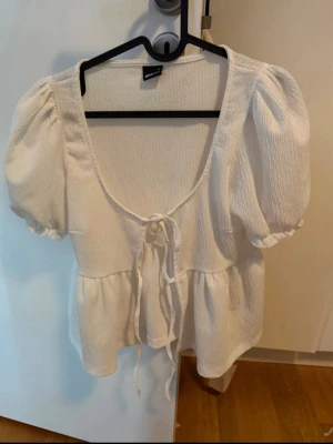 Vit blus med puffärm och knyt - Supersöt vit blus med puffiga korta ärmar och knyt framtill. Blusen har en djup v-ringning och volang nedtill, vilket ger en romantisk vibe. Perfekt till jeans eller kjol för en trendig look.