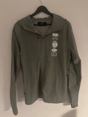 Windbreaker - Snygg grå vindjacka med huva och dragkedja framtill. Jackan har flera vita tryck på bröstet och en minimalistisk design. Tillverkad i ett lätt material som passar perfekt för blåsiga dagar. Långa ärmar och enkel passform.