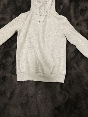 Ljusgrå basic hoodie med huva - En stilren ljusgrå hoodie med huva och dragsnören. Hoodien har långa ärmar, ribbade muddar och en klassisk känguruficka framtill. Perfekt för en avslappnad och enkel look. Materialet känns mjukt och bekvämt.