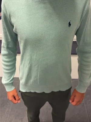 Mintgrön stickad tröja Polo Ralph Lauren - Snygg mintgrön stickad tröja från Polo Ralph Lauren i mjuk pima cotton. Tröjan har lång ärm, rund halsringning och den klassiska broderade loggan på bröstet. Perfekt för en stilren och avslappnad look.