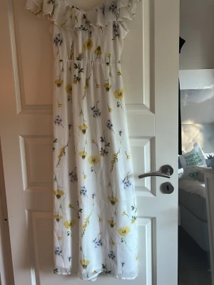 Vit långklänning med blommigt mönster - Söt vit långklänning med blommigt mönster i gult och blått. Klänningen har volang vid axlarna, offshoulder-modell och knyt i ryggen. Perfekt för sommarens alla tillfällen.