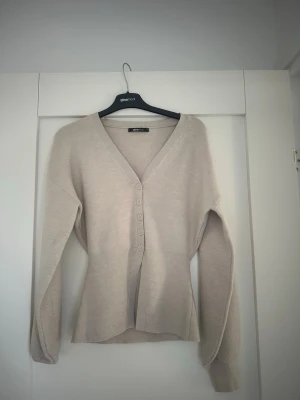 Beige kofta med v-ringning och knappar - Stilren beige kofta med v-ringning och fem knappar framtill. Modellen har lång ärm, ribbade muddar och markerad midja för en snygg siluett. Perfekt att slänga över en topp, t-shirt eller bara använda så. Materialet är mjukt och lättstickat. Aldrig använd då den bara legat i min garderob. 