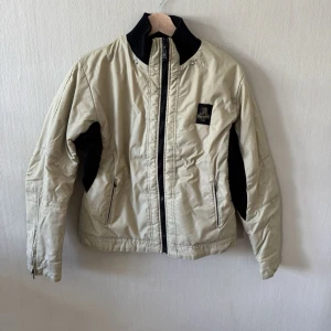 Refrigiwear Beige vindjacka med svarta detaljer - Stilren beige vindjacka med svarta paneler under armarna och svart hög krage. Jackan har dragkedja framtill, två sidofickor med dragkedja och en liten logopatch på bröstet. Perfekt för blåsiga dagar och enkel att matcha med olika outfits.0178