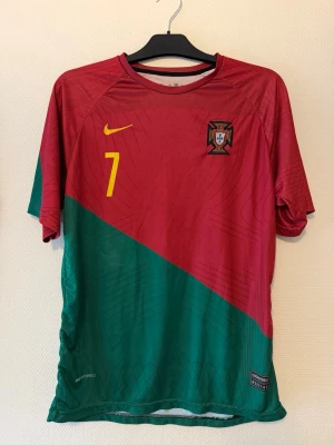 Portugal fotbollströja Ronaldo #7 - Snygg Portugal landslagströja från Nike med Ronaldo och nummer 7 på ryggen. Tröjan är röd och grön med diagonal färgdelning, gula detaljer och landslagets emblem på bröstet. Tillverkad i lätt och ventilerande material, perfekt för fotboll eller träning.