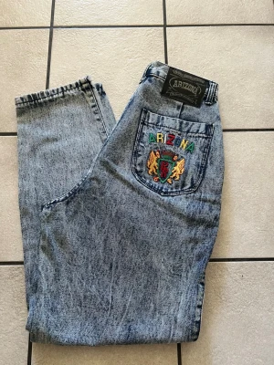 Arizona acid wash jeans med brodyr storlek 30 - Säljer ett par vintage-inspirerade acid wash jeans från Arizona med hög midja och raka ben. Bakfickan har färgglad brodyr med texten 'ARIZONA' och två lejon. Klassisk femficksmodell i blått denim, perfekt för dig som gillar retrostil. Finns ett litet håll, vänligen se bild.