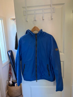 Blå och grå vändbar vindjacka colmar - Snygg vändbar vindjacka i blått och grått med huva och dragkedja framtill. Jackan har två sidofickor med dragkedja och en rund patch på ärmen. Perfekt för dig som vill ha två stilar i en jacka. Materialet är tunt och lätt, troligen polyester.