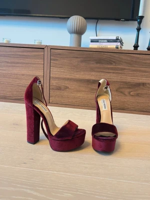 Steve Madden  - Snygga vinröda pumps från Steve Madden i sammetsmaterial. Skorna har hög blockklack, platåsula och öppen tå med bred rem över foten samt justerbar ankelrem. Perfekta för dig som vill sticka ut med en trendig och elegant look.