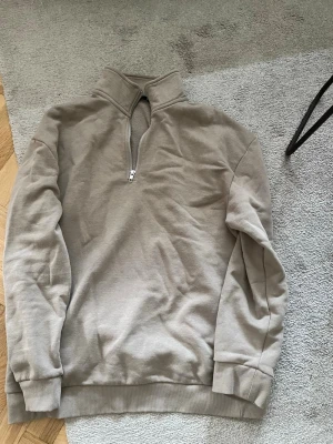 Beige hoodie med halv dragkedja - Säljer en beige hoodie med halv dragkedja framtill och hög krage. Hoodien har långa ärmar, ribbade muddar och är tillverkad i mjukt material. Perfekt för en avslappnad och stilren look.