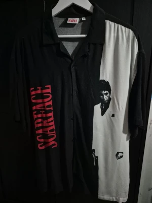 Scarface skjorta  - Unik svart och vit skjorta med Scarface-tryck och stor röd logga på framsidan. Skjortan har knappar framtill och korta ärmar. Perfekt för dig som gillar filmklassiker och vill sticka ut med en cool design.