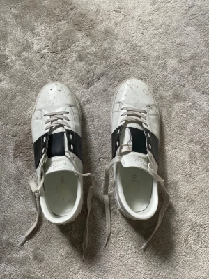 Valentino garavani - Snygga vita sneakers med bred svart rem över mitten och vita skosnören. Skorna har en låg profil och är tillverkade i läder med gummisula. Perfekta för en clean och stilren look.