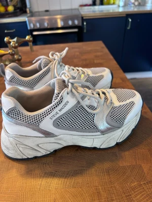 Chunky vita sneakers Steve Madden st 38 - Chunky sneakers från Steve Madden i vitt med silvriga detaljer och meshpaneler för extra ventilation. Skorna har tjock sula och snörning framtill. Perfekta för dig som gillar en sportig och trendig look.