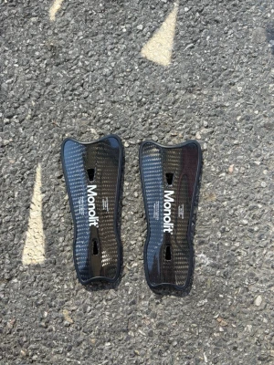 Monolit benskydd i kolfiber - Säljer ett par svarta benskydd från Monolit i kolfiber. De har en snygg glansig finish och är lätta med ergonomisk form. Perfekta för dig som vill ha extra skydd och stil på planen. Texten 'Carbon Shinguards' och 'Equilateral fit' står på framsidan.