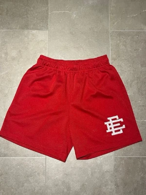 Eric Emanuel shorts  - Säljer ett par röda mesh shorts med elastisk midja och en vit broderad bokstavsdetalj på vänster ben. Shortsen har en loose passform och är perfekta för sommarens alla aktiviteter.
