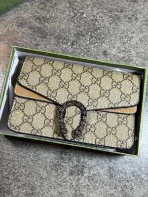 Gucci Axel väska  - Snygg beige kuvertväska  Gucci med klassiskt GG-monogram över hela väskan. Väskan har en dekorativ metallspänne framtill och svarta detaljer längs kanterna. Tillverkad i canvas med läderdetaljer. Perfekt för dig som gillar lyxiga accessoarer. Kom dm för diskutera pris 