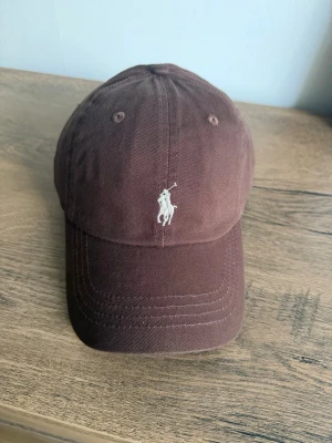 Brun keps från Polo Ralph Lauren - Snygg brun keps från Polo Ralph Lauren med den klassiska broderade loggan framtill. Justerbar baktill för perfekt passform. Passar både dam och herr. Perfekt till vardags eller sommarens aktiviteter!