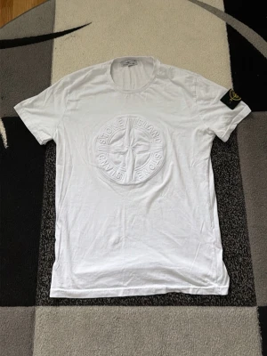 Vit Stone Island t-shirt med emblem - Vit t-shirt från Stone Island med stort broderat emblem på bröstet och klassisk logga på ärmen. T-shirten har rund hals och korta ärmar, perfekt för en clean och stilren look.
