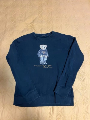 Marinblå sweatshirt Polo Ralph Lauren - Säljer en marinblå sweatshirt från Polo Ralph Lauren med den ikoniska björnen på bröstet. Tröjan har rund hals, långa ärmar och ribbade muddar. Perfekt för dig som gillar klassisk streetwear med en twist.