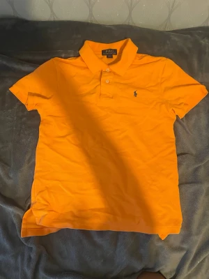 Orange pikétröja från Polo Ralph Lauren - Snygg orange pikétröja från Polo Ralph Lauren med klassisk krage, korta ärmar och knappslå framtill. Tröjan har den ikoniska lilla loggan broderad på bröstet och är tillverkad i mjuk bomull. Perfekt för en avslappnad och stilren look. Kan passa 12-13 också