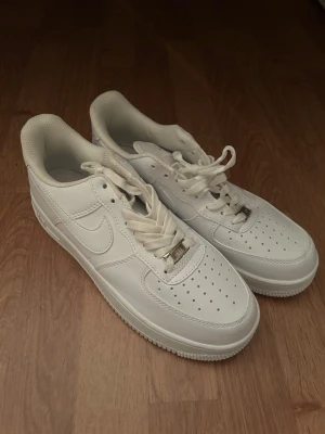 Nike Air Force 1 vita sneakers - Klassiska Nike Air Force 1 sneakers i helvitt läder med perforerad tå, vita skosnören och diskret Nike-logga på sidan. Snygg och tidlös modell med låg profil och robust sula. Perfekta för dig som gillar en clean och stilren look.