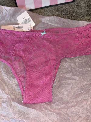 Rosa spets-trosa med rosett - Säljer en snygg rosa trosa i spets med blommönster och en liten ljusblå rosett framtill. Trosan har låg midja och är tillverkad i ett tunt, stretchigt material som känns bekvämt mot huden. Perfekt för dig som gillar feminina detaljer.