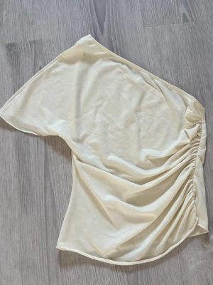 Krämvit oneshoulder topp - Snygg krämvit oneshoulder topp med draperad detalj på sidan. Toppen har en asymmetrisk design och är tillverkad i ett mjukt, tunt material som känns skönt mot huden. 