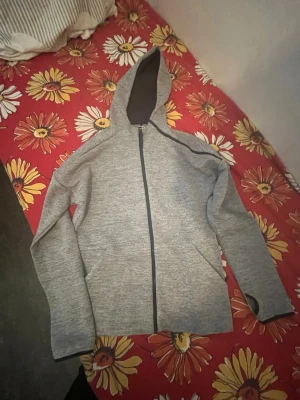 Grå hoodie med dragkedja från Adidas för kids 176 cm - Snygg gråmelerad hoodie från Adidas med hel dragkedja och huva. Tröjan har svarta detaljer, tumhål i ärmsluten och två sidofickor. Tillverkad i mjukt material som känns skönt mot huden. Perfekt för chill eller träning.