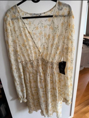 Blommig klänning från NA-KD - Söt klänning från NA-KD i ljus, krämvit ton med gult blommönster. Klänningen har v-ringning, långärmade ärmar med resår vid handlederna och smock i midjan för en bekväm passform. 