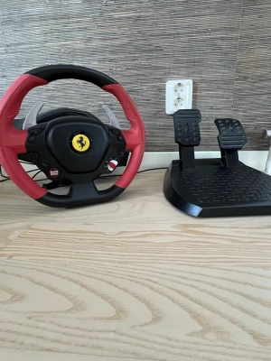 Ratt och pedaler Ferrari-design - Säljer en gamingratt och pedalset med Ferrari-design. Ratten är svart och röd med Ferrari-logga i mitten och har växlingspaddlar i silver. Pedalerna är svarta och har halkskyddad yta. Perfekt för racing på konsol eller PC.