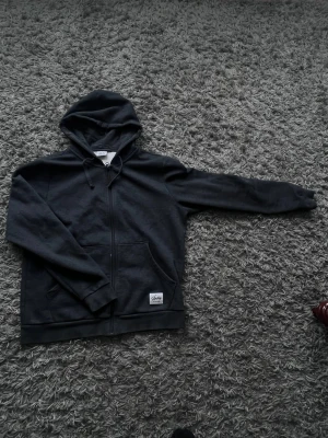 Mörkgrå zip hoodie med huva - Säljer en mörkgrå hoodie med dragkedja och huva. Hoodien har fickor framtill och en liten vit patch nertill med text. Perfekt för en avslappnad och stilren look. Materialet känns mjukt och bekvämt.