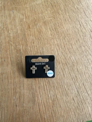 Korsformade örhängen från Montini - Snygga korsformade örhängen i guldton från Montini. Örhängena är dekorerade med små glittrande stenar som ger ett elegant intryck. Perfekta för dig som vill ha en trendig accessoar med lite edge.