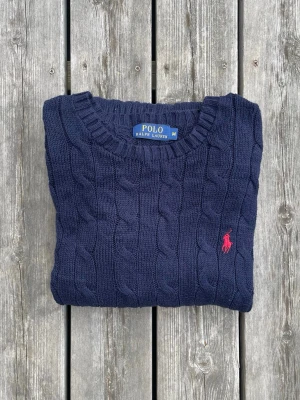  Polo Ralph Lauren Kabelstickad | Marinblå - Strl M - Kabelstickad mörkblå tröja från Polo Ralph Lauren med rund halsringning och ikonisk röd broderad logga på bröstet. Tröjan har ribbade muddar och är gjord i mjukt material som passar perfekt till höst och vinter. Fint använt skick! Skickas alltid samma dag eller dagen efter