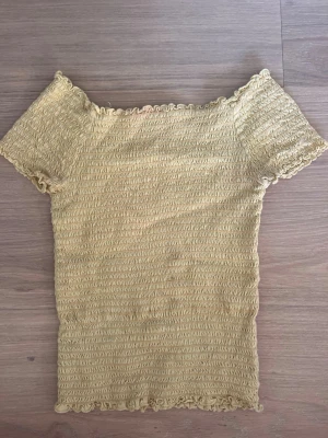 Gul smockad topp  - Säljer en ljusgul t-shirt med smockat typ. T-shirten har korta ärmar och vågiga kanter vid hals, ärmslut och nederkant. Storlek 146/152 men funkar till större