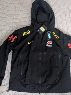 Svart Brasilien vindjacka Nike - Snygg svart vindjacka från Nike med Brasilien-tema. Jackan har huva, dragkedja framtill och flera loggor i gult, rött och grönt. Märken som 'vivo', 'itau' och Brasil-logga syns tydligt. Perfekt för dig som gillar sportig stil och streetwear.