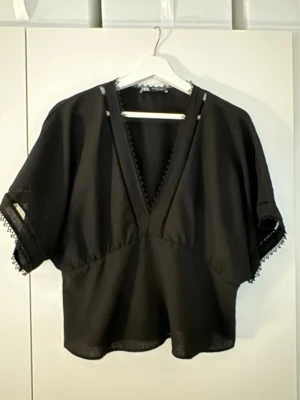 Svart blus med spetsdetaljer - Snygg svart blus med v-ringning och korta, vida ärmar. Blusen har fina spetsdetaljer längs ärmslut och halsringning som ger en unik touch. Perfekt för dig som gillar en avslappnad men ändå stilren look.