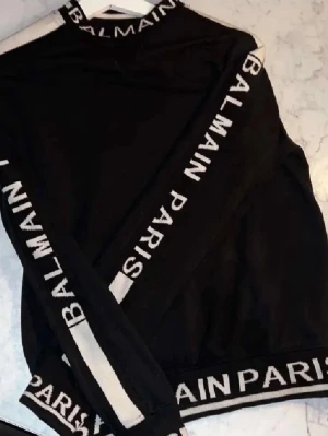 Svart Balmain Paris sweatshirt - Svart sweatshirt från Balmain Paris med vita detaljer och logotyp längs krage, ärmar och nederkant. Tröjan har rund hals, långa ärmar och en sportig look. Perfekt för dig som gillar streetwear och exklusiva märken.