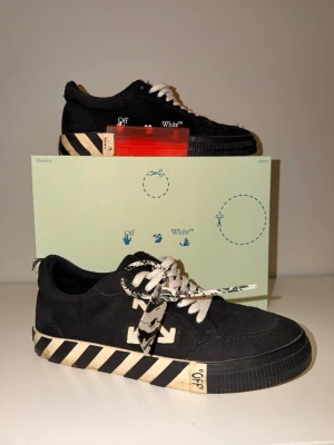 Off-White svarta låga sneakers - Svarta sneakers från Off-White med vita detaljer och signaturpilar på sidan. Skorna har vita snören med svart text, randig sula i svart och vitt samt en röd tag på sidan. Tillverkade i canvas och gummi. Box medföljer.