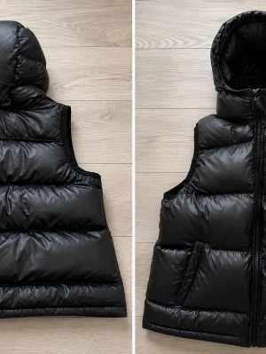 Svart dunväst med huva - Snygg svart dunväst med huva och dragkedja framtill. Västen har två fickor på sidorna och breda, quiltade paneler som ger en puffig look. Perfekt för lager-på-lager och streetstyle. Materialet är glansigt och ger en modern vibe.