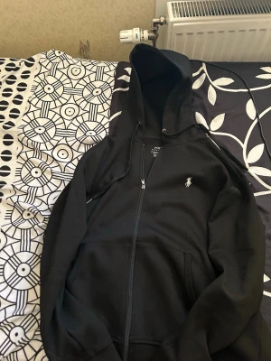 Svart hoodie från Polo Ralph Lauren - Säljer en svart hoodie från Polo Ralph Lauren med dragkedja och huva. Klassisk logga broderad på bröstet i vitt. Hoodien har fickor framtill och justerbar dragsko i huvan. Perfekt för en chill och stilren look.