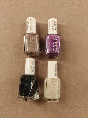 Fyra nagellack i olika färger - Säljer fyra nagellack i olika färger: beige, lila, svart och ett genomskinligt med glitter. Lacken är från Essie och har används max 2 gånger 