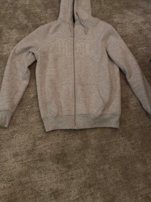 Tuttolente hoodie - Snygg grå zip hoodie med huva och fickor framtill. På bröstet finns broderade bokstäver 'M' och 'T' i vitt. Hoodien har ribbade muddar vid ärmslut och nederkant. Perfekt för en avslappnad och trendig stil.
