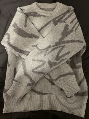 Grå stickad tröja med abstrakt mönster - Säljer en stickad tröja i ljusgrått med mörkgrått abstrakt mönster. Tröjan har rund halsringning, ribbade muddar och nederkant. Perfekt för dig som gillar en chill och modern stil.