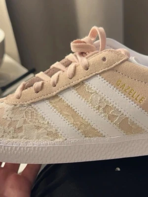Adidas Gazelle spetsdetaljer beige - Snygga Adidas Gazelle sneakers i beige mocka med dit satta vita detaljer och spetsmönster pa ovandelen. Ar hemma gjord och har inga defekter på skorna mer än den tillsatta spetsen. Klassiska tre rander på sidan och vit sula. Skorna har rosa skosnören och gulddetaljer på sidan och plösen. Perfekta för dig som vill ha en unik touch på dina sneakers. Spetsen som är tillsatt har hålt mycket bra och finns inga täcken på slitage, men dem är hemma gjorde så man har det i å tanke.