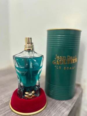 Jean Paul Gaultier Le Beau parfym - Unik parfymflaska formad som en manlig torso i blått glas med gulddetaljer. Kommer med en elegant grön metalltub med guldfärgad text och står på en röd sammetskudde. Perfekt för dig som gillar snygg design och ikoniska dofter.